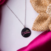 A sterling silver pendant chain with a round pendant engraved with the word 'Princess' presented on a pink fabric surface.